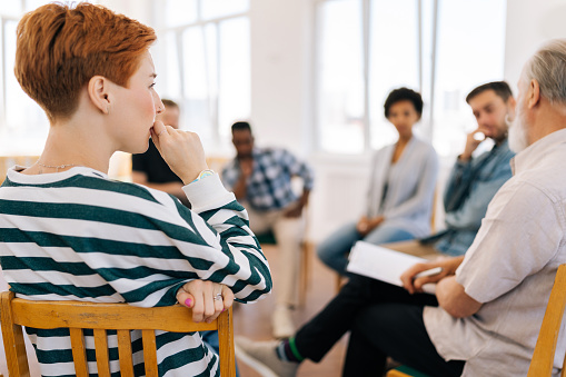 Rear view of thoughtful young woman thinking about mental problem or addiction sitting in circle during group therapy session. Concept of mental health, psychotherapy, depression, social issues.